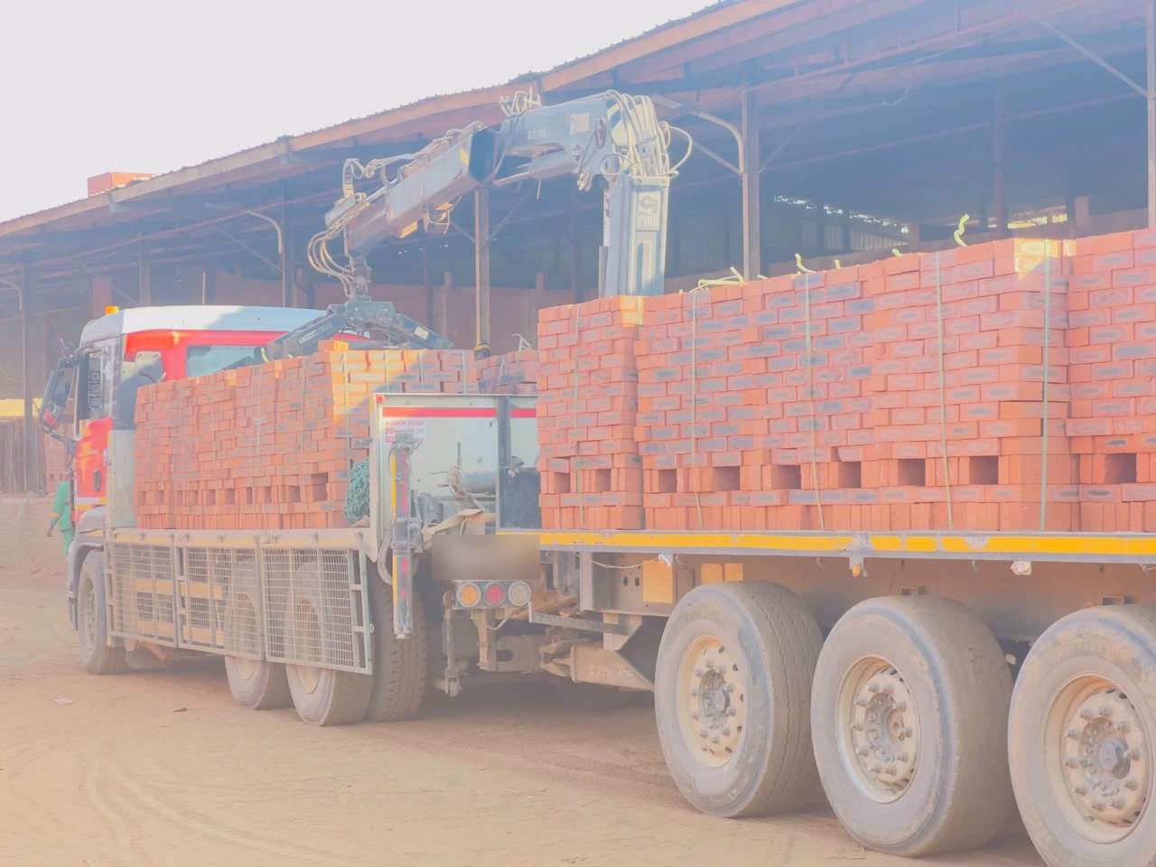 A crane truck loaded end to end with pallets of red face brick under the drying sheds in Ruwa