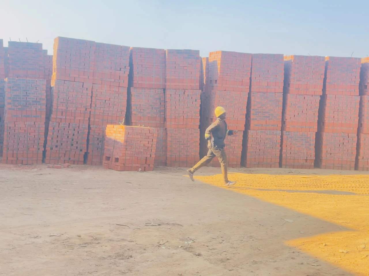 A worker in a hard hat walking past a long wall of brick pallets at the Ruwa site